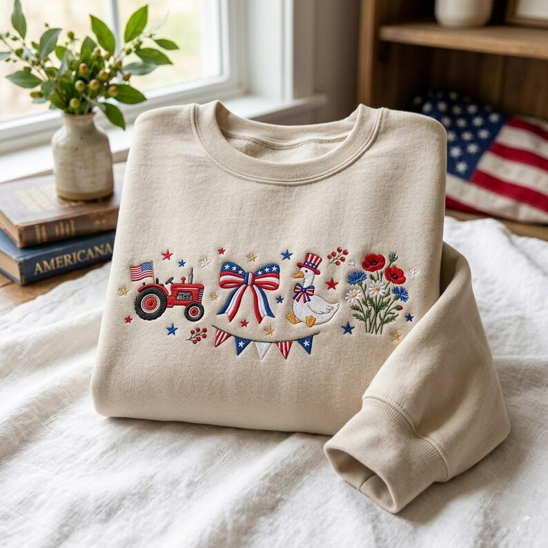 K&ouml;nnte beinhalten: Beiges Sweatshirt mit gesticktem patriotischem Design. Das Design umfasst einen roten Traktor, eine Schleife mit rot-wei&szlig;-blauen Streifen, eine Gans mit Zylinder und verschiedene Blumen und Sterne. Das Sweatshirt hat einen Rundhalsausschnitt und lange &Auml;rmel.
