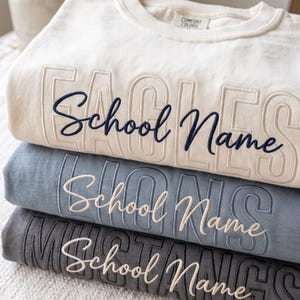 May include: A stack of three crewneck sweatshirts in cream, dusty blue, and charcoal gray. Each features embossed block lettering of school mascots and the embroidered script text School Name. The top sweatshirt has a Comfort Colors label.