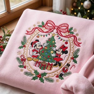 May include: A light pink sweatshirt featuring an embroidered design of Mickey and Minnie Mouse decorating a Christmas tree. The scene is framed by a gold scalloped border, a red and white gingham bow, holly leaves, candy canes, and a nutcracker.