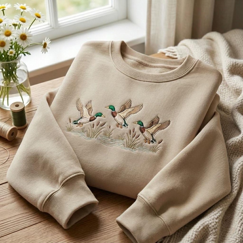 Puede incluir: Sudadera beige de cuello redondo con un dise&ntilde;o bordado de tres patos volando sobre juncos y agua. Los patos tienen cabezas verdes, alas marrones y detalles azules. La sudadera est&aacute; doblada sobre una superficie de madera.
