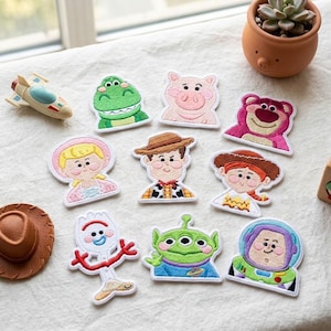 Può includere: Una collezione di dieci toppe ricamate raffiguranti vari personaggi dei cartoni animati, tra cui un dinosauro verde, un maiale rosa, un orso magenta, una persona bionda, un cowboy, una cowgirl, una forchetta bianca, un alieno verde e un astronauta.