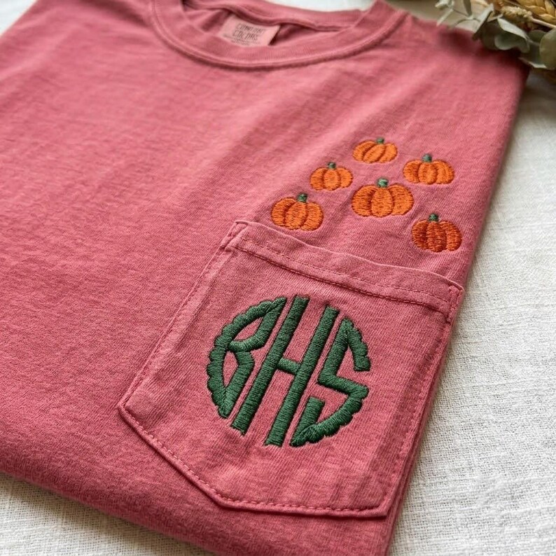 Może przedstawiać: T-shirt w kolorze przybrudzonego r&oacute;żu z kieszonką na piersi, na kt&oacute;rej wyhaftowano zielony monogram z literami BHS. Nad kieszonką znajduje się wyhaftowany wz&oacute;r składający się z sześciu małych pomarańczowych dyń ułożonych w grupę.
