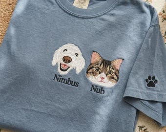 Può includere: Una t-shirt blu a maniche corte con ritratti ricamati di un cane bianco e di un gatto tigrato marrone. Il testo "Nimbus" è cucito sotto il cane e "Nub" sotto il gatto. Un'impronta di zampa nera è ricamata sulla manica, con etichetta Comfort Colors.