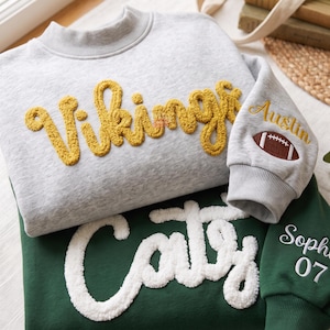Puede incluir: Una sudadera gris claro con letras de felpa amarillas que dicen Vikings y un parche de balón de fútbol americano en la manga con el nombre Austin. Debajo, una sudadera verde oscuro con letras de felpa blancas que dicen Caty y una manga con el nombre Sophie y el número 07.