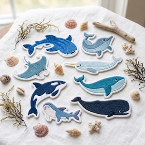 May include: A collection of eight embroidered fabric patches featuring various marine animals including a whale shark, dolphin, manta ray, narwhal, orca, and several whales, arranged on a white fabric surface with scattered seashells and driftwood.