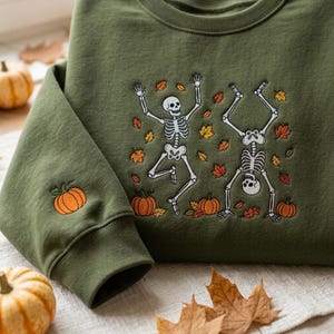 Op de afbeelding: Een olijfgroene sweater met ronde hals voorzien van een borduursel van twee witte skeletten die dansen tussen verspreide oranje en gele herfstbladeren en kleine pompoenen. Een enkele oranje pompoen is geborduurd op de boord van de linkermouw.