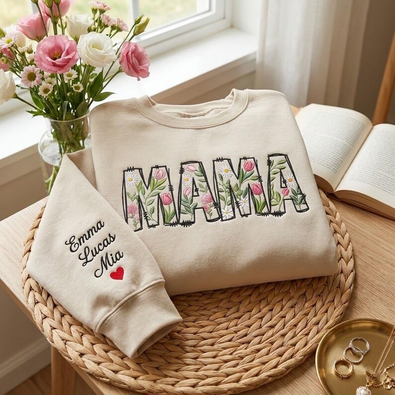 Op de afbeelding: Beige sweatshirt met het woord "MAMA" geborduurd met een bloemenontwerp. De mouw heeft de namen "Emma Lucas Mia" en een rood hart. De sweatshirt ligt op een geweven mat.