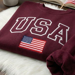 Może przedstawiać: Bordowa bluza z napisem "USA" w białych literach. Poniżej znajduje się haftowana flaga amerykańska. Bluza jest złożona na białej, puszystej powierzchni.