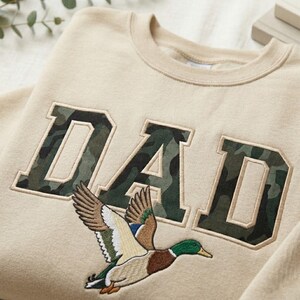 May include: Beige sweatshirt with the word "DAD" in camouflage-patterned letters. A detailed embroidered image of a flying duck is below the letters. The sweatshirt has a crew neck and a relaxed fit, suitable for casual wear.