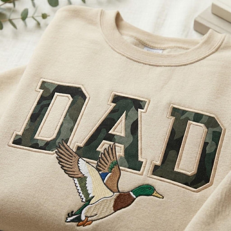 May include: Beige sweatshirt with the word "DAD" in camouflage-patterned letters. A detailed embroidered image of a flying duck is below the letters. The sweatshirt has a crew neck and a relaxed fit, suitable for casual wear.