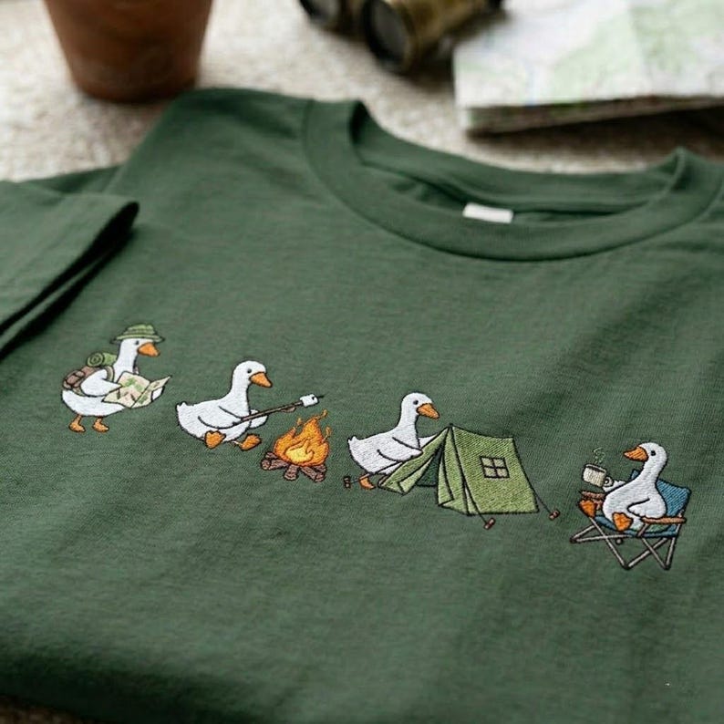 Op de afbeelding: Groen t-shirt met een geborduurd ontwerp van kamperende ganzen. E&eacute;n gans draagt een hoed en rugzak, een andere roostert een marshmallow, een derde staat bij een tent en de laatste nipt uit een mok in een stoel.