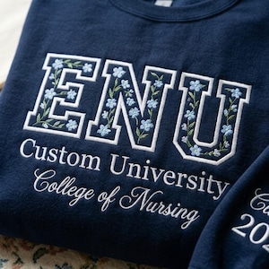 May include: Navy blue sweatshirt with white embroidered text and floral details. The word "ENU" is prominently displayed, filled with light blue flowers and green leaves. Below, the text reads "Custom University College of Nursing".