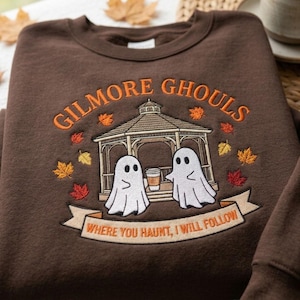 May include: A dark brown crewneck sweatshirt featuring an embroidered design of a gazebo with two white ghosts holding a coffee cup. The text reads GILMORE GHOULS and WHERE YOU HAUNT, I WILL FOLLOW, surrounded by small orange, red, and yellow autumn leaves.