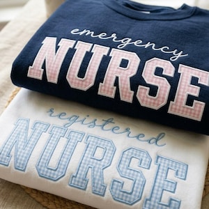 May include: Two sweatshirts, one navy blue and one white, are stacked. The navy blue sweatshirt has the words "emergency NURSE" in white script and pink gingham letters. The white sweatshirt says "registered NURSE" in light blue gingham letters.
