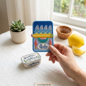May include: A rectangular embroidered patch featuring a blue background with four stylized fish, a yellow horizontal bar, and the text "Quite THE CATCH" in white and blue. Below it sits a small ceramic box decorated with blue fish illustrations.
