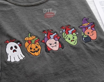 Może przedstawiać: Ciemnoszary t-shirt z pięcioma wyhaftowanymi wzorami anatomicznych serc w stylu halloweenowym. Postacie obejmują białego ducha, pomarańczową dynię, wampira, zielonego potwora w stylu Frankensteina oraz czarownicę w fioletowym kapeluszu.