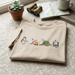 Op de afbeelding: Beige T-shirt met een geborduurd ontwerp van kamperende cartoon ganzen. E&eacute;n gans draagt een kaart, een andere roostert een marshmallow en een andere zit in een kampeerstoel. Het shirt heeft een ronde hals en korte mouwen.