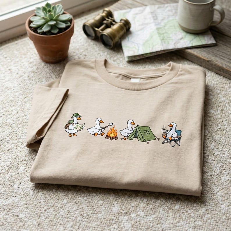 Op de afbeelding: Beige T-shirt met een geborduurd ontwerp van kamperende cartoon ganzen. E&eacute;n gans draagt een kaart, een andere roostert een marshmallow en een andere zit in een kampeerstoel. Het shirt heeft een ronde hals en korte mouwen.