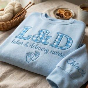 Pode incluir: Camisola azul clara com bordados. As letras "L&D" estão num padrão xadrez azul e branco, com "labor & delivery nurse" por baixo. A manga tem o nome "Chloe" bordado em itálico.