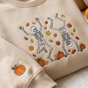 Op de afbeelding: Een beige sweater met ronde hals voorzien van een geborduurd ontwerp van twee witte skeletten die dansen tussen verspreide oranje, gele en bruine herfstbladeren en oranje pompoenen. Een enkele oranje pompoen is geborduurd op de mouwboord.
