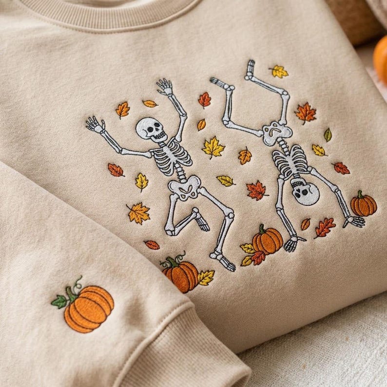 Op de afbeelding: Een beige sweater met ronde hals voorzien van een geborduurd ontwerp van twee witte skeletten die dansen tussen verspreide oranje, gele en bruine herfstbladeren en oranje pompoenen. Een enkele oranje pompoen is geborduurd op de mouwboord.