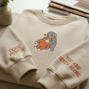 May include: Beige sweatshirt with embroidered cartoon monkeys hugging, a red heart above them. The left sleeve cuff reads "TEAM PUNCH" in red, and the right sleeve cuff reads "YOU ARE NEVER ALONE" in red.