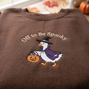 May include: A dark brown crewneck sweatshirt featuring an embroidered design of a white duck wearing a purple cape and a black witch hat. The duck holds an orange jack-o'-lantern candy bucket. The text Off to Be Spooky is embroidered above.