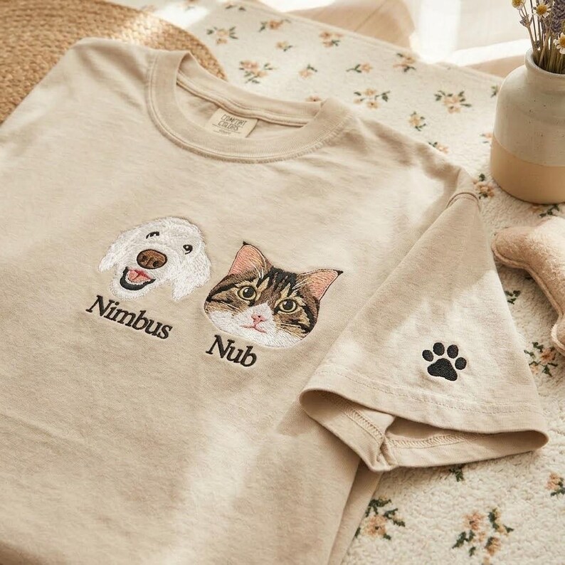 May include: A beige t-shirt featuring embroidered portraits of a white dog and a brown and white tabby cat. The name Nimbus is stitched below the dog, and the name Nub is stitched below the cat. A small black paw print is embroidered on the sleeve.