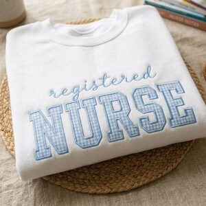 May include: A white sweatshirt with the words "registered NURSE" embroidered in light blue. The word "registered" is in a cursive font, and "NURSE" is in a block letter style with a blue and white gingham pattern.