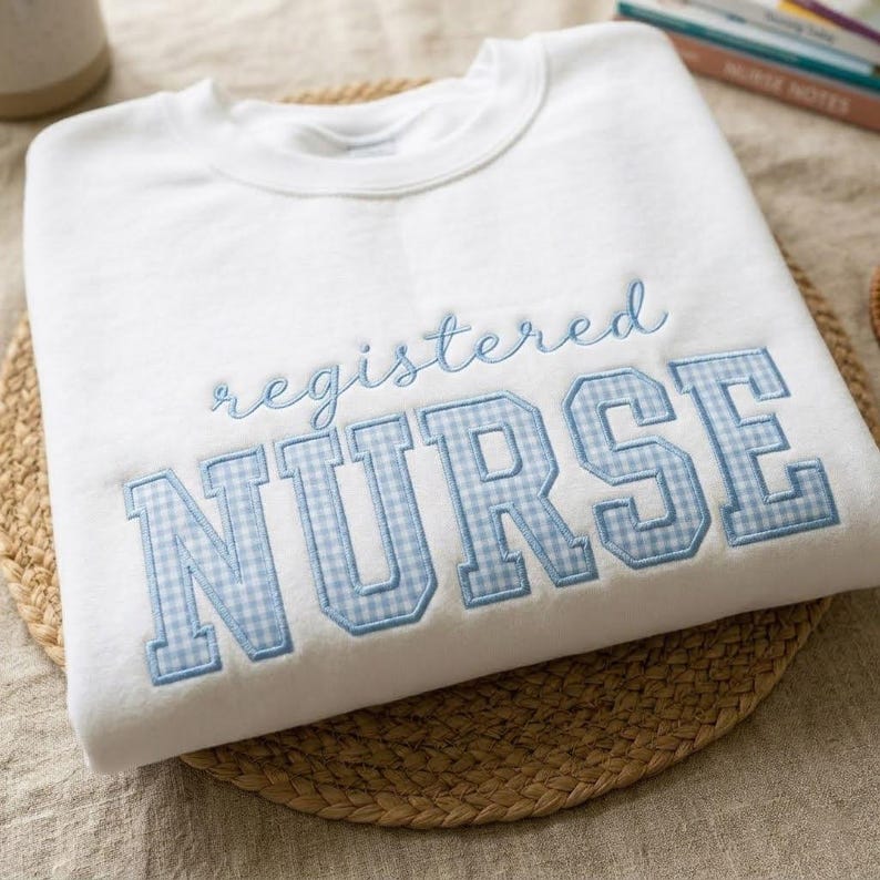 May include: A white sweatshirt with the words "registered NURSE" embroidered in light blue. The word "registered" is in a cursive font, and "NURSE" is in a block letter style with a blue and white gingham pattern.