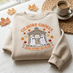 May include: A beige crewneck sweatshirt featuring an embroidered design of a gazebo with two ghosts holding a coffee cup. The text reads GILMORE GHOULS and WHERE YOU HAUNT, I WILL FOLLOW, surrounded by small orange, yellow, and red embroidered autumn leaves.