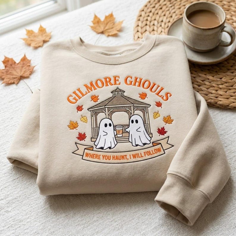 May include: A beige crewneck sweatshirt featuring an embroidered design of a gazebo with two ghosts holding a coffee cup. The text reads GILMORE GHOULS and WHERE YOU HAUNT, I WILL FOLLOW, surrounded by small orange, yellow, and red embroidered autumn leaves.