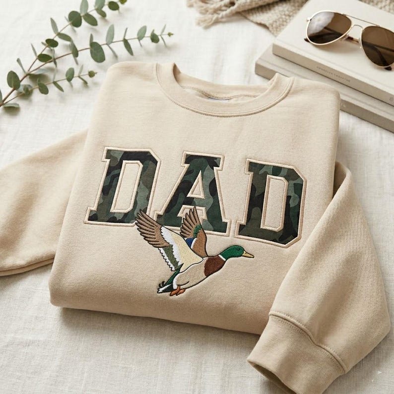 May include: Beige sweatshirt with the word "DAD" in camouflage-style lettering. A detailed embroidered mallard duck is flying below the text. The sweatshirt has a crew neck and long sleeves.