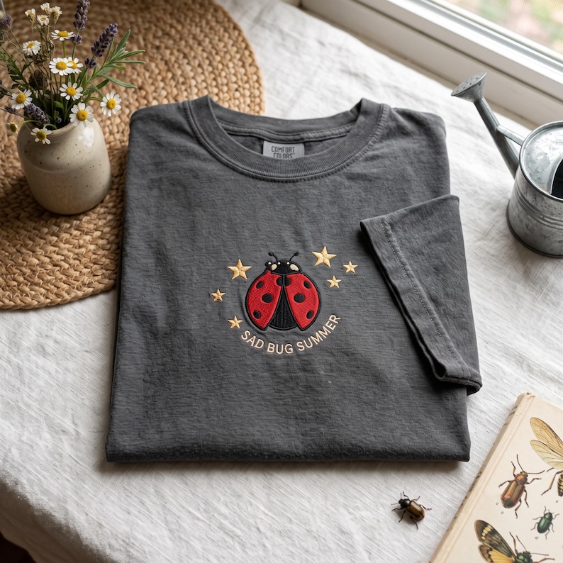 May include: A charcoal gray short-sleeve t-shirt featuring an embroidered red and black ladybug surrounded by small yellow stars. Below the ladybug, the text reads "SAD BUG SUMMER" in a simple, light-colored font. The shirt has a crew neckline.