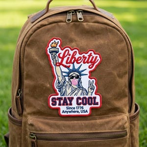 Pode incluir: Uma mochila de lona castanha com um grande emblema bordado da Estátua da Liberdade a usar óculos de sol e a fazer uma bolha cor-de-rosa. O emblema inclui o texto Liberty, STAY COOL e Since 1776 Anywhere, USA em letras vermelhas e azuis.