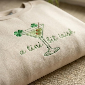 Könnte beinhalten: Beiges Sweatshirt mit grünem Stickdesign. Das Design zeigt ein Martini-Glas mit Oliven und Kleeblättern sowie den Text "a tini bit irish". Das Sweatshirt ist gefaltet.