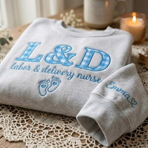May include: A gray sweatshirt and matching hat with blue embroidery. The sweatshirt features "L&D labor & delivery nurse" with blue gingham letters and baby feet. The hat is embroidered with the name "Emma".