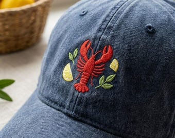 Peut inclure: Une casquette de baseball en jean bleu marine avec un motif brodé sur le panneau avant. La broderie représente un homard rouge vif centré entre deux quartiers de citron jaunes et deux petites feuilles vertes, cousus avec des fils détaillés.
