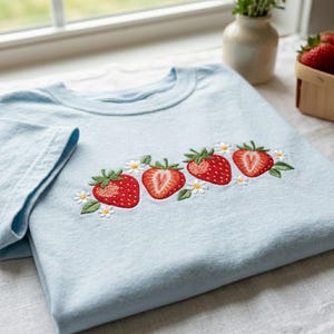 May include: A light blue t-shirt featuring an embroidered design across the chest. The embroidery depicts a horizontal row of four red strawberries, including two whole berries and two halved berries, interspersed with small white daisy flowers.