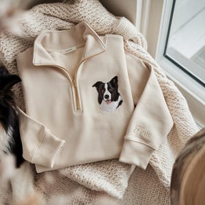 May include: A cream-colored sweatshirt with a gold zipper and a black and white dog embroidered on the front. The name "Finn" is embroidered on the sleeve. The sweatshirt is folded on a cream-colored blanket.