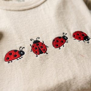 May include: A beige t-shirt featuring four embroidered ladybugs arranged in a horizontal row across the chest. Each ladybug is depicted with a bright red shell, black spots, and black antennae, showcasing detailed stitching on the fabric.
