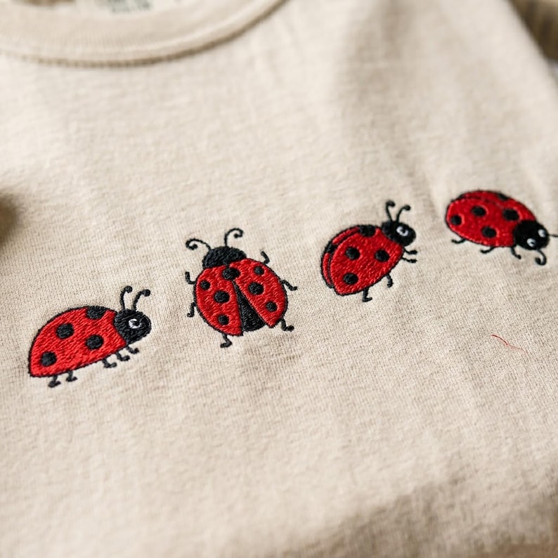 May include: A beige t-shirt featuring four embroidered ladybugs arranged in a horizontal row across the chest. Each ladybug is depicted with a bright red shell, black spots, and black antennae, showcasing detailed stitching on the fabric.