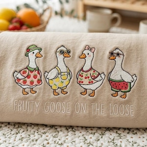 Pode incluir: Tecido bege bordado com quatro gansos de desenho animado usando roupas com tema de frutas e óculos de sol. Os gansos estão vestidos com desenhos de morango, limão, cereja e melancia. O texto "FRUITY GOOSE ON THE LOOSE" é bordado abaixo.