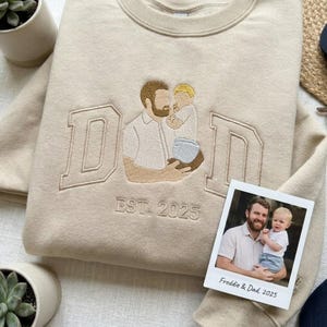 May include: Beige sweatshirt with embroidered "DAD" lettering and a father-child illustration. The sweatshirt includes "EST. 2025" and a small photo with the text "Freddie & Dad, 2025". The design is in shades of beige and brown.