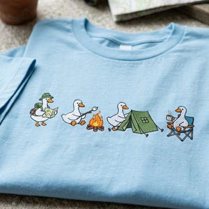 Op de afbeelding: Lichtblauw T-shirt met een geborduurd ontwerp van kamperende ganzen. E&eacute;n gans draagt een kaart, een andere roostert een marshmallow, een derde zet een tent op en de laatste drinkt koffie in een campingstoel. Het ontwerp is in een grillige stijl.