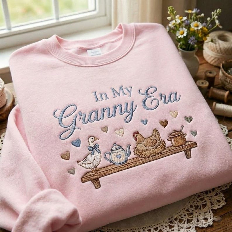 May include: A light pink sweatshirt with the embroidered phrase "In My Granny Era." Below the text is a shelf with a duck, teapot, chicken, and pot. The design includes heart shapes.