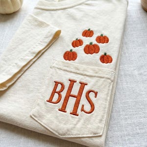 Może przedstawiać: Kremowy t-shirt z wyhaftowaną kieszonką na piersi z pomarańczowymi literami BHS. Nad kieszonką wyhaftowano grupę sześciu małych pomarańczowych dyń z zielonymi ogonkami na materiale koszulki.