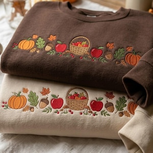 May include: Two crewneck sweatshirts, one dark brown and one cream, each featuring an embroidered horizontal band of autumn motifs including orange pumpkins, red apples, a woven basket, brown acorns, green oak leaves, and small mushrooms.