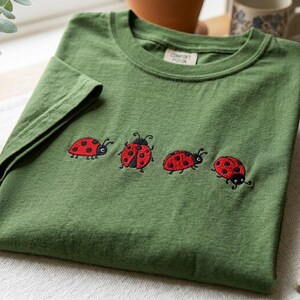 May include: An olive green short-sleeved t-shirt featuring four embroidered ladybugs in red and black. The ladybugs are positioned in a horizontal row across the chest. A white interior neck label displays the text Comfort Poton in black ink.