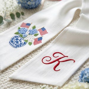 May include: White linen kitchen towels. One towel features an embroidered blue and white vase with blue flowers and American flags. The other towel has a red embroidered letter "K".